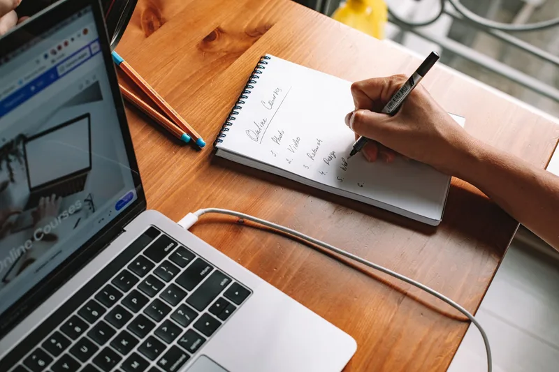 Person writing notes in a notebook while studying an online course on a laptop at home