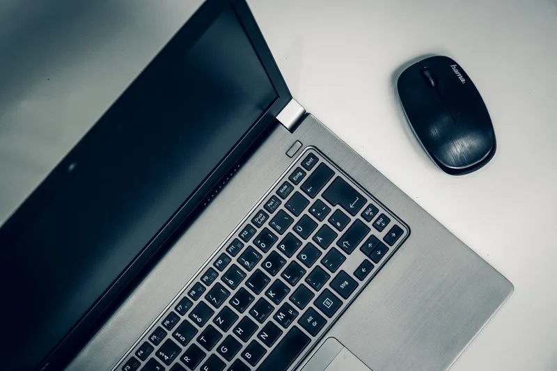 A plain laptop and simple wireless mouse on a desk, representing a modest, affordable home workspace setup for a beginner virtual assistant