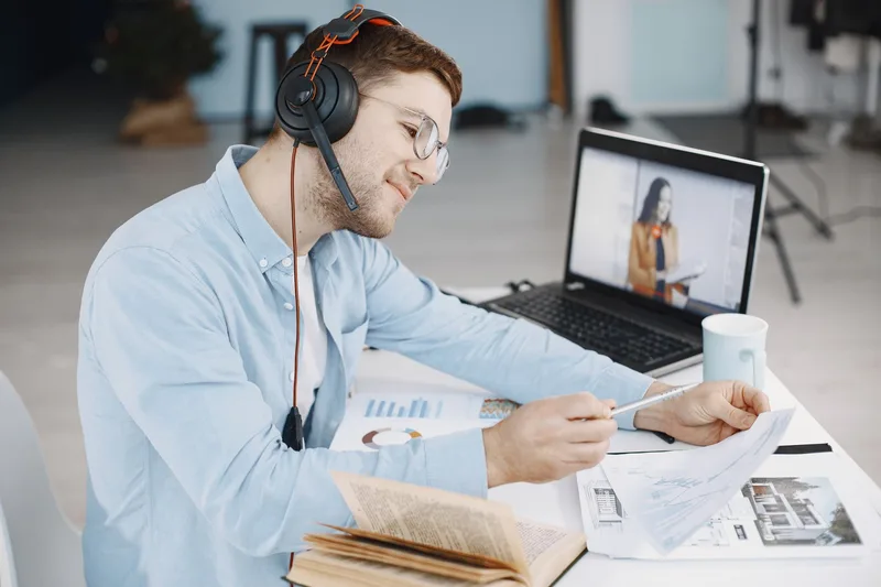 Man working remotely with headphones, reviewing documents, and video conferencing on laptop