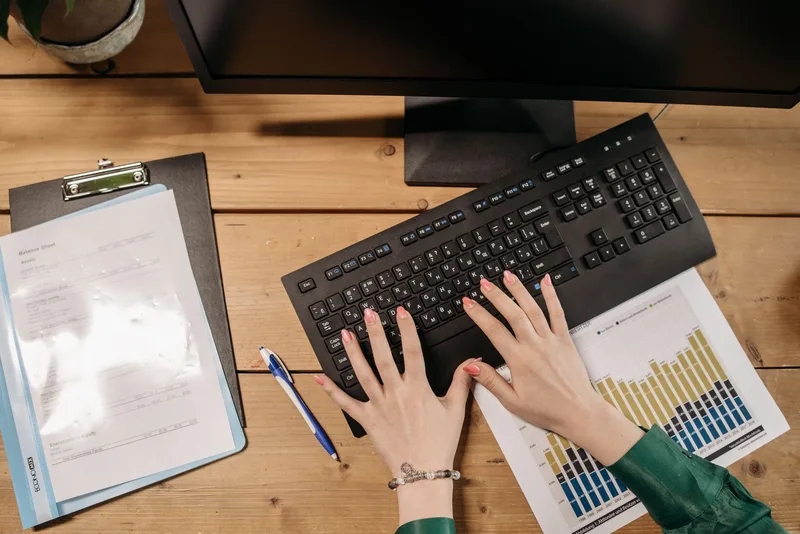 Hands typing on a keyboard next to printed spreadsheets and charts on a desk