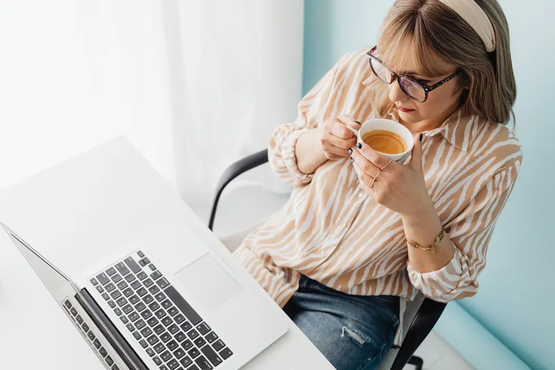 Woman in glasses sipping coffee while looking at an open laptop at a white desk