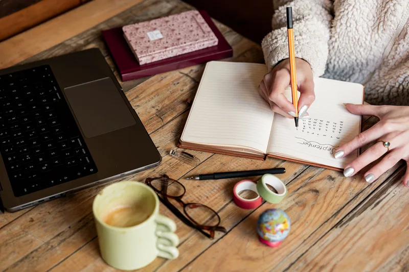 Person writing a daily plan in a notebook beside a laptop and a mug of coffee on a wooden desk